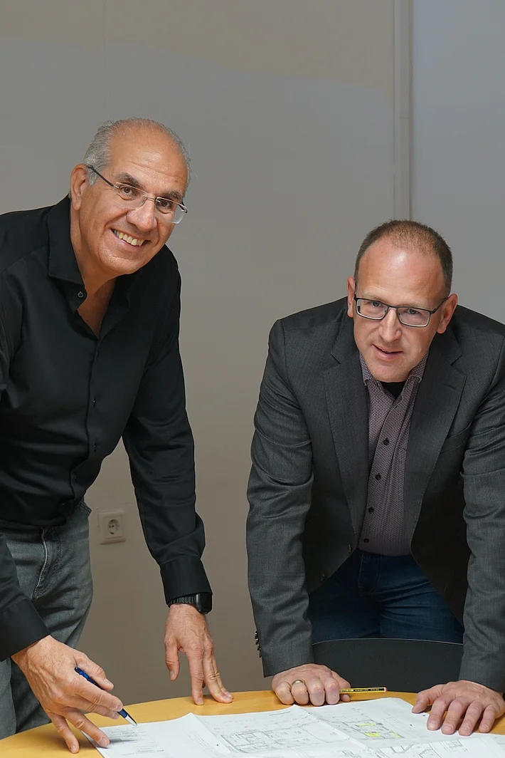 Bereichsleiter Josef Pfeiffer (links im Bild) und Werkhof‑Geschäftsführer Peter Astashenko stehen an einem Tisch, betrachten einen Plan und schauen in die Kamera.