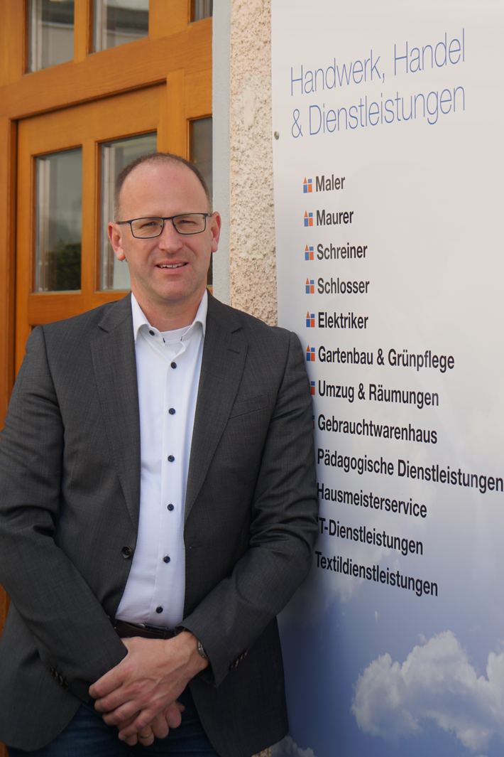 Geschäftsführer Peter Astaschenko lehnt an der Außenwand des Werkhof Regensburg, im Hintergrund ist ein Schild mit den Gewerken des Werkhofs teilweise sichtbar.