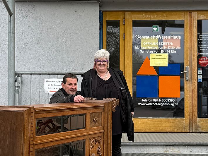 Zwei Mitarbeitende des Gebrauchtwarenhauses Regensburg stehen vor dem Eingang des Gebäudes. Auf einem Rollwagen vor ihnen befindet sich ein gerade angelieferter Schrank zur Warenannahme.