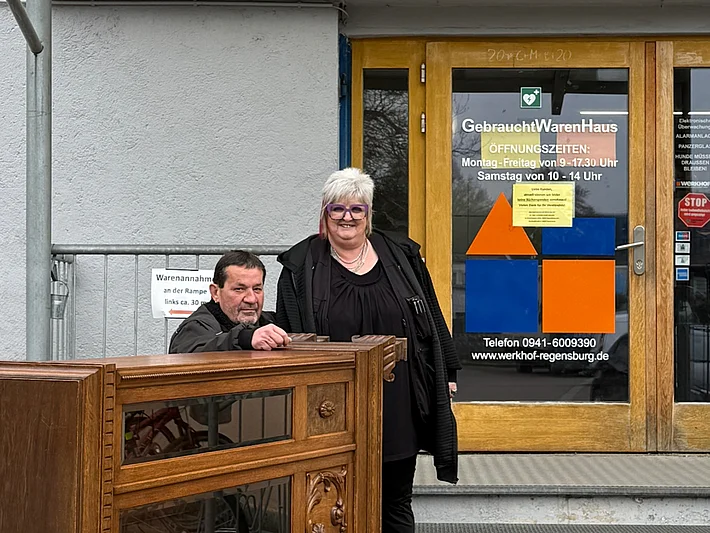Zwei Mitarbeitende des Gebrauchtwarenhauses Regensburg stehen vor dem Eingang des Gebäudes. Auf einem Rollwagen vor ihnen befindet sich ein gerade angelieferter Schrank zur Warenannahme.