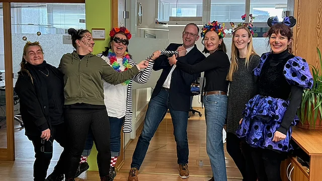 Sechs leicht verkleidete Mitarbeiterinnen des Werkhof Regensburg stehen gemeinsam mit Geschäftsführer Peter Astashenko in einem Raum. Sie lächeln in die Kamera. Dem Geschäftsführer wird die Krawatte abgeschnitten . als Zeichen der Weiberfaschings-Tradition.